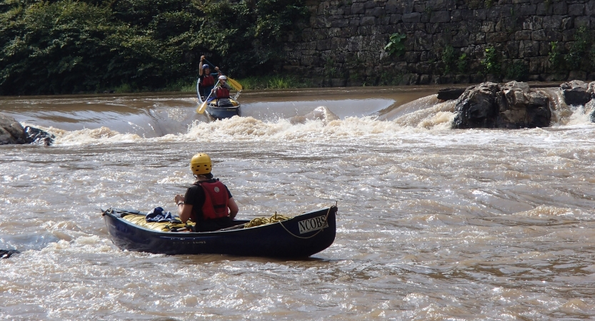 One person wearing safety gear sits in a canoe, watching two other people wearing safety gear paddle a canoe over a whitewater rapid.